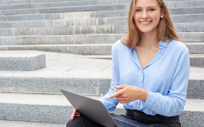Eine junge Frau sitzt auf Stufen, sie hat einen Laptop auf dem Schoß und lächelt