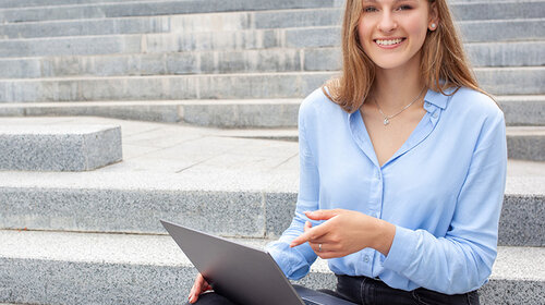 Eine junge Frau sitzt auf Stufen, sie hat einen Laptop auf dem Schoß und lächelt